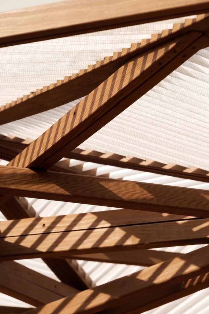 Close-up of intersecting wooden beams with sunlight casting striped shadows on a white ceiling surface.