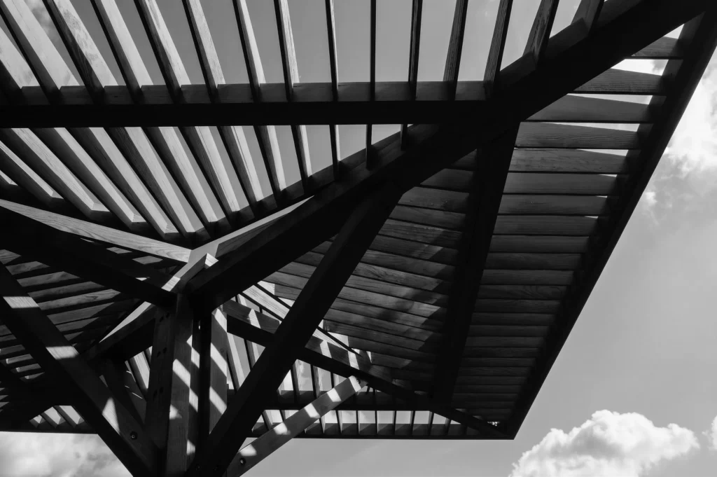 Contemporary wooden canopy structure casting geometric shadows through layered slats against an open sky, illustrating the interaction between light, shade, and architecture.